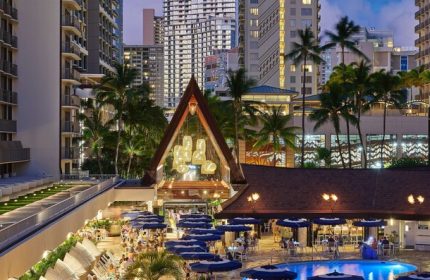 Outrigger Reef Waikiki Beach Resort pool and dining area at dusk. Gay-friendly hotel with palm trees and city skyline. Waikiki travel.