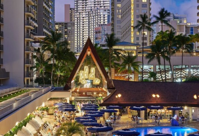 Outrigger Reef Waikiki Beach Resort pool and dining area at dusk. Gay-friendly hotel with palm trees and city skyline. Waikiki travel.