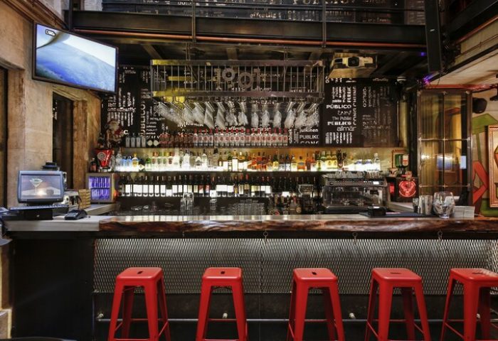 Hotel The Public bar. Gay-friendly hotel interior with red stools, wood bar, liquor bottles, and TV. Special Category hotel for gay men travel.
