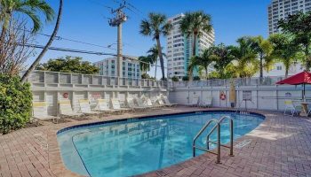 Pool at The Grand Resort and Spa, an all-male, gay-friendly hotel. Lounge chairs line the pool's edge. Palm trees and buildings in the background.