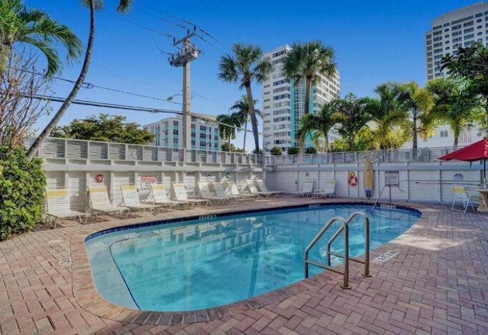 Pool at The Grand Resort and Spa, an all-male, gay-friendly hotel. Lounge chairs line the pool's edge. Palm trees and buildings in the background.