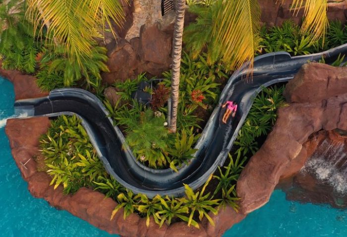 The Royal Hawaiian, a Luxury Collection Resort, Waikiki gay-friendly hotel. Person on water slide at pool. Tropical plants and rock formations.