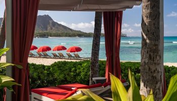 Sheraton Waikiki Beach Resort view of Diamond Head from a cabana. Red umbrellas and lounge chairs on the beach. Gay-friendly hotel.