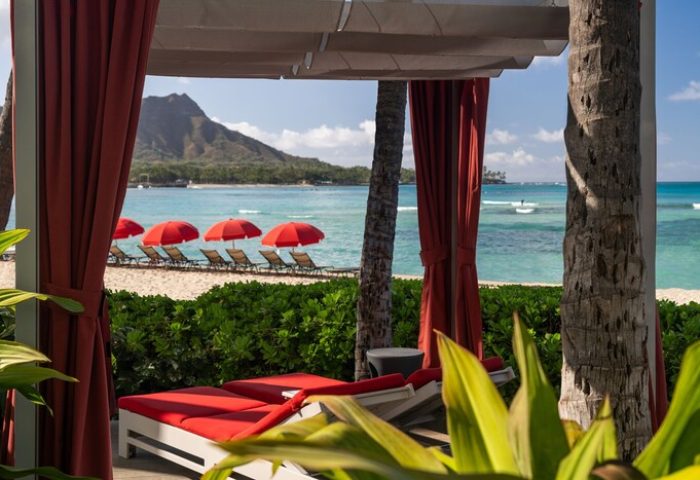 Sheraton Waikiki Beach Resort view of Diamond Head from a cabana. Red umbrellas and lounge chairs on the beach. Gay-friendly hotel.