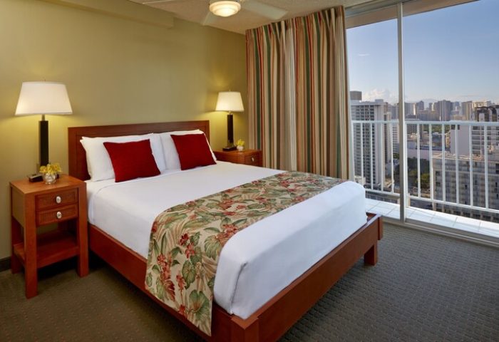 Pacific Monarch Hotel room showcasing a bed with red pillows, tropical runner, and city view. Gay-friendly hotel with balcony.