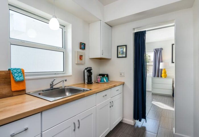Nobleton Hotel's gay-friendly kitchenette. White cabinets, wood countertop, sink, & coffee maker. Bedroom view w/ blue curtain. Modern design.