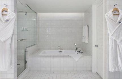 Halekulani Hotel bathroom with white tile, shower, and tub. Bathrobes hang nearby. Gay-friendly hotel offers luxury travel for gay men.