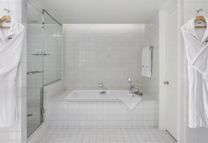 Halekulani Hotel bathroom with white tile, shower, and tub. Bathrobes hang nearby. Gay-friendly hotel offers luxury travel for gay men.