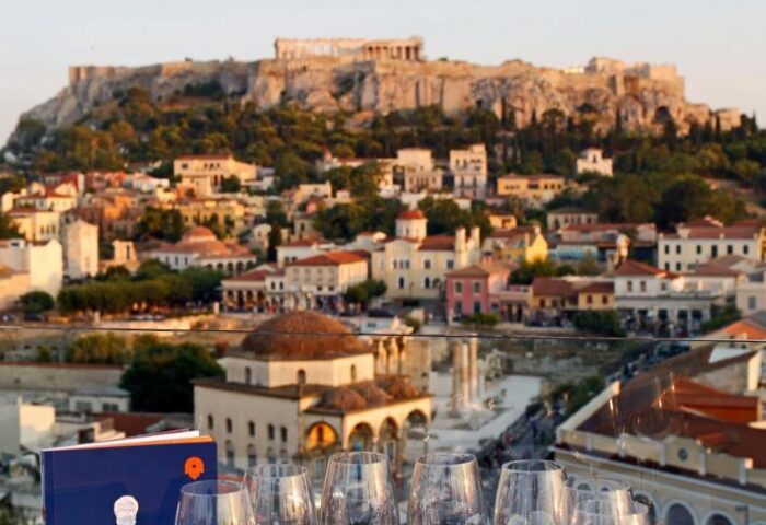 A For Athens Hotel view. Wine glasses, Acropolis backdrop. Gay-friendly hotel offers wine tasting with iconic Athens scenery. Greece travel.