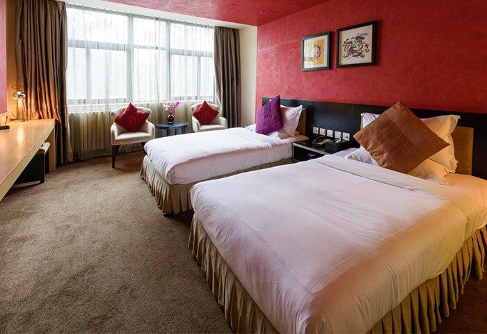 Gay-Friendly Hotel A. HOTEL: Twin beds in a room with red walls, desk, window, and two chairs. Comfortable lodging for gay men travelers.