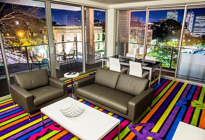 ADGE Apartments, a gay-friendly hotel: Interior view of a modern apartment with colorful striped rug, city view, and balcony at night.