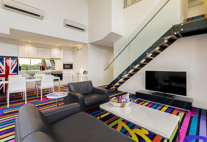 ADGE Apartments gay-friendly hotel room. Modern interior with colorful striped carpet, kitchen, staircase, and Union Jack fridge.