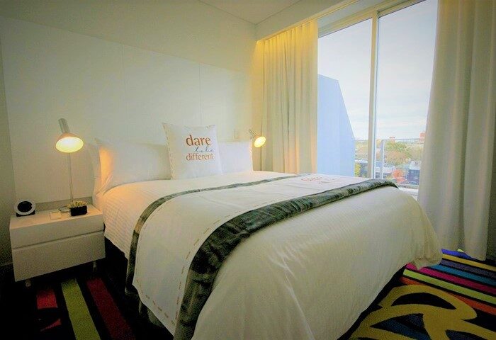 ADGE Apartments gay-friendly hotel room. A bed with a dare to be different pillow, bedside table, lamp, and a colorful rug. Window view.