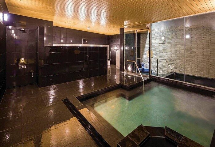 APA Hotel Shinjuku Gyoemmae's onsen. This gay-friendly hotel in Japan features a traditional Japanese bath with dark tile and a wooden ceiling.