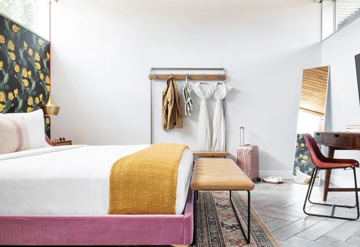 ARRIVE Palm Springs gay-friendly hotel room. Pink bed frame, white linens, yellow throw, cactus wallpaper, desk, and luggage. Stylish gay travel.