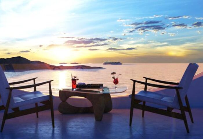 Mykonos sunset view from Absolute Mykonos Suites & More, a gay-friendly hotel. Cocktails on table, chairs, sea, cruise ship. Perfect gay travel.