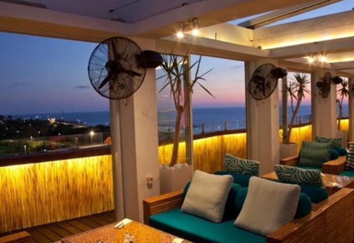 Alexander Tel Aviv Hotel's gay-friendly terrace at dusk. Sea view seating area with fans, plants, and cozy cushions. Perfect for gay travelers.