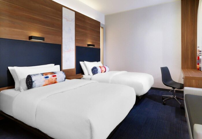 Aloft Munich: Gay-friendly hotel room with two twin beds, desk, and chair. Modern design with wood paneling and colorful pillows. Comfy for gay travelers.