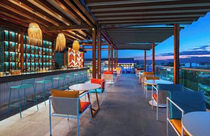 Aloft Playa del Carmen gay-friendly hotel rooftop bar at dusk. Tables, chairs, and barstools. Bottles on shelves behind the bar. Modern design.