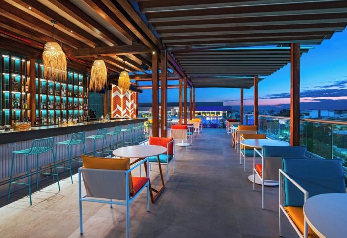 Aloft Playa del Carmen gay-friendly hotel rooftop bar at dusk. Tables, chairs, and barstools. Bottles on shelves behind the bar. Modern design.