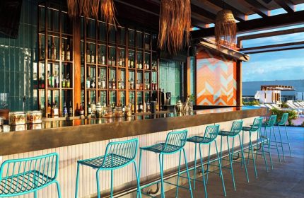 Aloft Playa del Carmen's bar area, a gay-friendly hotel. Blue chairs line the bar, with bottles displayed behind. Outdoor pool visible.