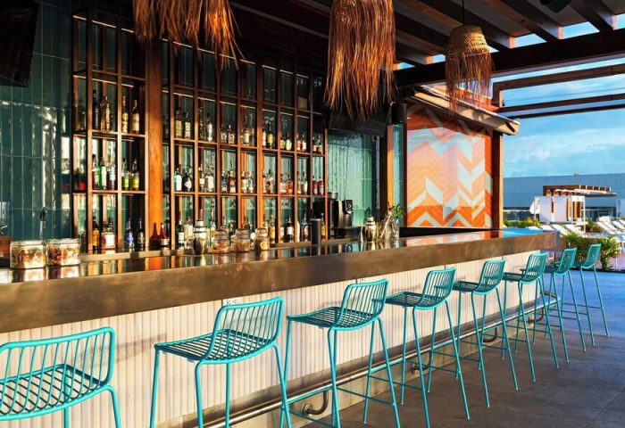Aloft Playa del Carmen's bar area, a gay-friendly hotel. Blue chairs line the bar, with bottles displayed behind. Outdoor pool visible.