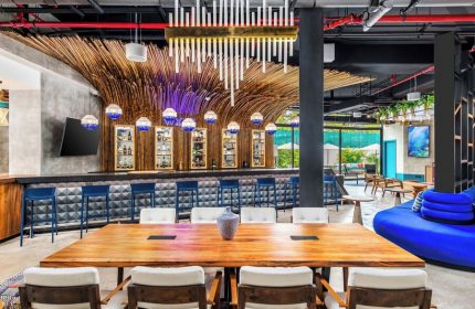 Aloft Tulum, a gay-friendly hotel: bar area with bamboo decor, blue stools, and a large wooden table. Outdoor pool view. Modern design.