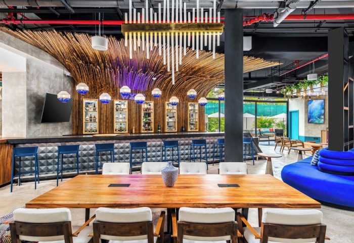 Aloft Tulum, a gay-friendly hotel: bar area with bamboo decor, blue stools, and a large wooden table. Outdoor pool view. Modern design.