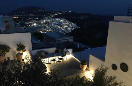 Amelot Art Suites, a gay-friendly hotel. Night view from a suite's terrace, showcasing a table, chairs, & city lights in the background.