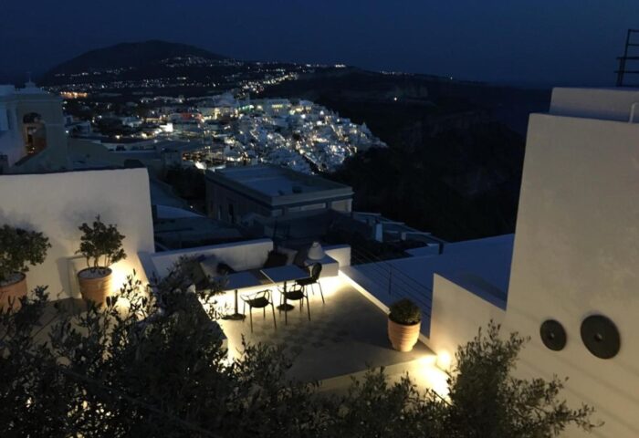 Amelot Art Suites, a gay-friendly hotel. Night view from a suite's terrace, showcasing a table, chairs, & city lights in the background.