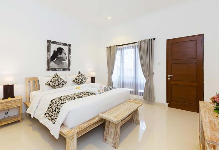Amerta Seminyak gay-friendly hotel room. Bedroom with bed, bench, bedside table, lamp, curtains, and picture. Gay travel advice in Bali.
