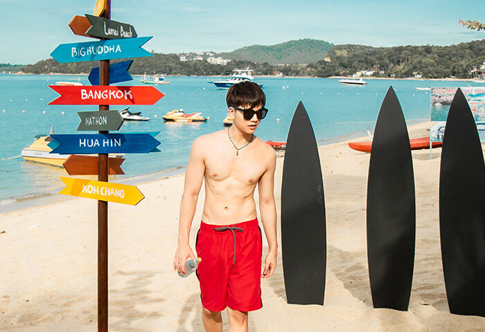 Gay-friendly Anantara Bophut Koh Samui Resort: Man on the beach with surfboards and destination signpost. Thailand travel for gay men.