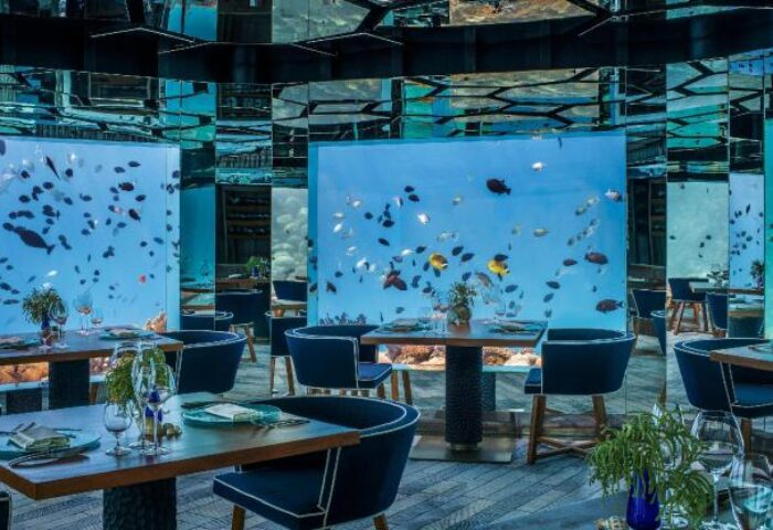 Gay-Friendly Hotel Anantara Kihavah Maldives Villas underwater restaurant. Tables and chairs surround aquarium walls with fish.