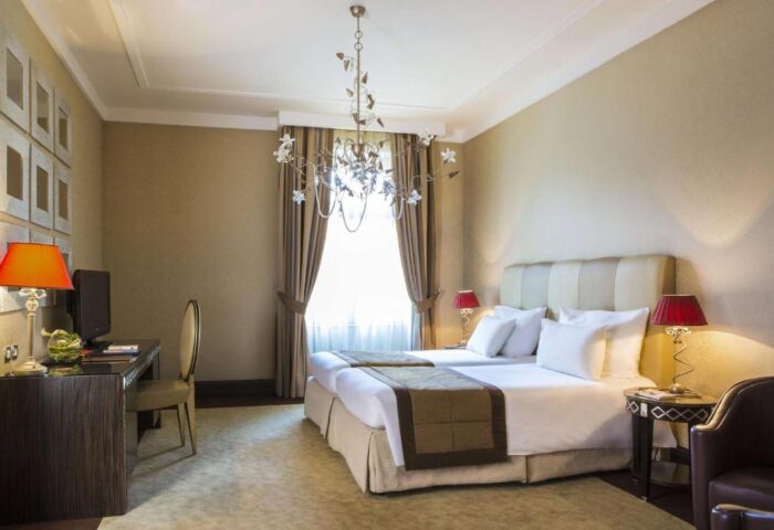 Anantara New York Palace Budapest room. Gay-friendly, pet-friendly hotel with twin beds, desk, chair, chandelier, and elegant decor.