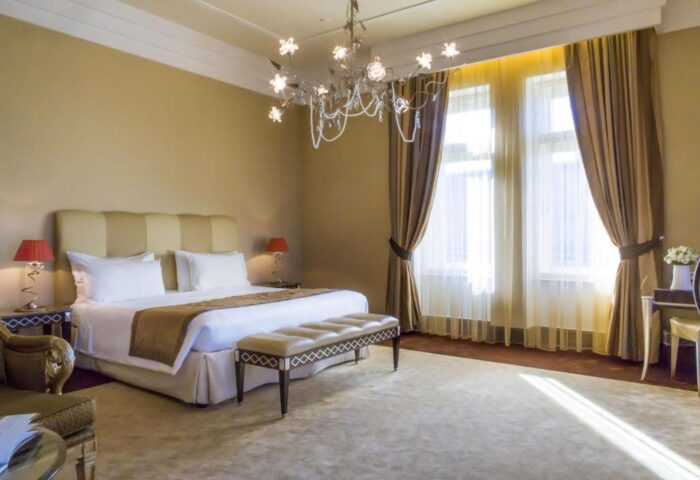 Anantara New York Palace Budapest bedroom. Gay-friendly, pet-friendly hotel room with king bed, chandelier, and large windows with curtains.