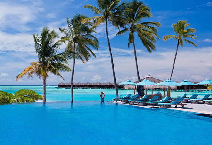 Infinity pool at gay-friendly Anantara Veli Maldives Resort. Palm trees, blue umbrellas, and overwater bungalows. Perfect for gay travel.