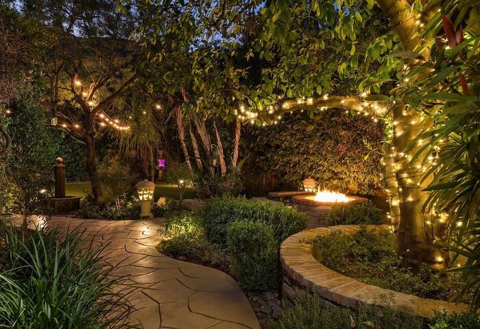 Gay-friendly Andalusian Court Hotel garden at night. A stone path leads to a fire pit surrounded by lush greenery and string lights.