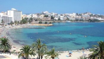 Gay-Friendly Hotel Apartamentos Lido view. Beach, blue water, palm trees, and buildings in Ibiza. Ideal for gay men travel advice.