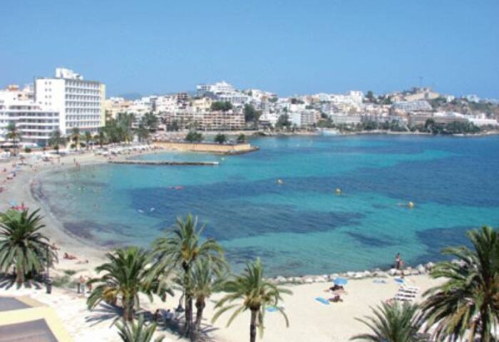 Gay-Friendly Hotel Apartamentos Lido view. Beach, blue water, palm trees, and buildings in Ibiza. Ideal for gay men travel advice.