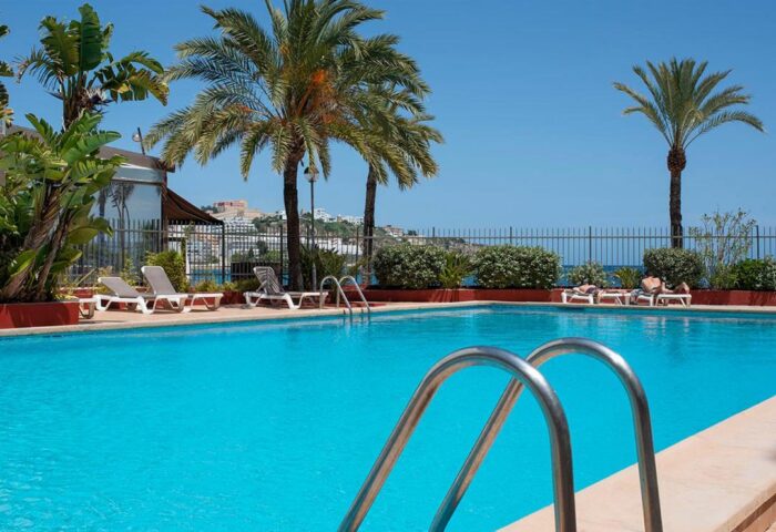 Apartamentos Llobet pool view. Gay-friendly hotel with sunbeds, palm trees, and clear blue water. Ideal for gay travel and relaxation.