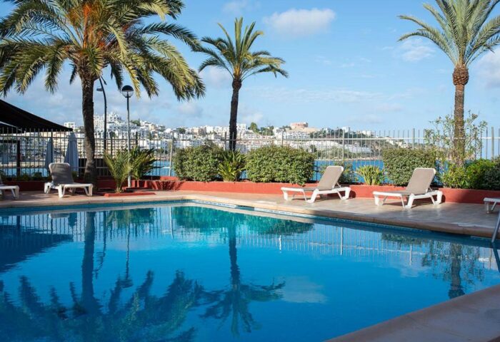 Apartamentos Llobet pool view. Gay-friendly hotel with palm trees, lounge chairs, and a scenic view of the city and sea.