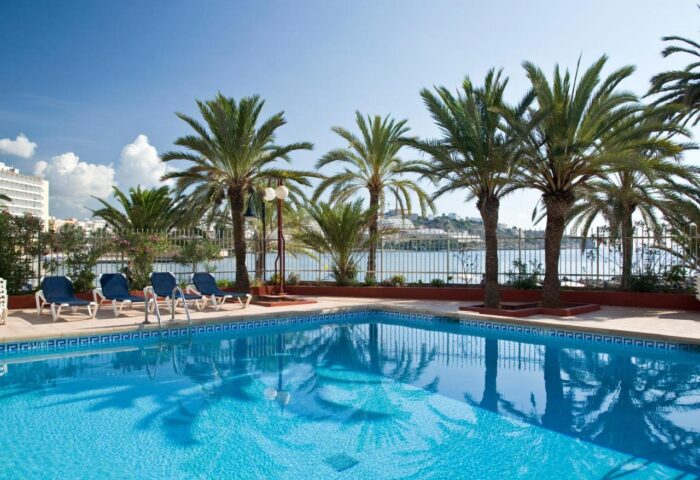 Apartamentos Llobet pool view. Gay-friendly hotel with palm trees, blue water, and lounge chairs. Perfect for gay travel and relaxation.