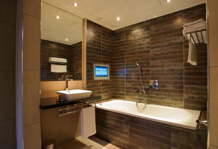 Bathroom in Roomzzz Aparthotel Manchester City, a gay-friendly hotel. Features a bathtub, sink, mirror, and TV. Modern design.