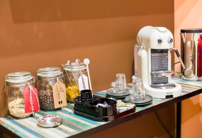 MarcoAurelio49 Apartments' gay-friendly hotel breakfast bar. Jars of oats, chocolate, pasta, coffee maker, cups, and pods on a colorful table.
