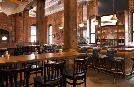 Argonaut Hotel's gay-friendly bar area. Brick walls, wooden beams, and nautical decor create a warm, inviting atmosphere. Bar stools and tables.