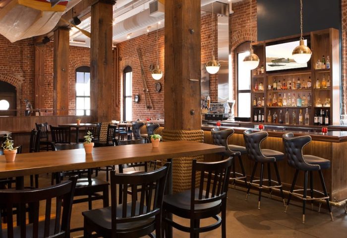 Argonaut Hotel's gay-friendly bar area. Brick walls, wooden beams, and nautical decor create a warm, inviting atmosphere. Bar stools and tables.