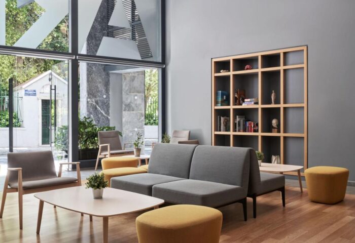 Arion Athens Hotel's lobby. Gay-friendly hotel seating area with gray sofas, wooden chairs, yellow ottomans, and a bookshelf. Modern design.
