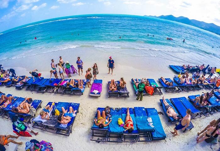 Gay-friendly Ark Bar Beach Resort: Beachgoers relax on blue lounge chairs on white sand. Turquoise water, mountains in the distance.