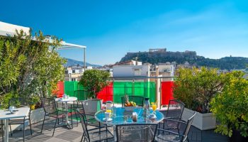 Attalos Hotel rooftop terrace view of Acropolis. Gay-friendly hotel in Athens, Greece. Outdoor seating and clear blue sky.