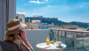 Woman on phone at Attalos Hotel, a gay-friendly hotel, with Acropolis view. Enjoying breakfast on balcony in Athens. Travel advice for gay men.